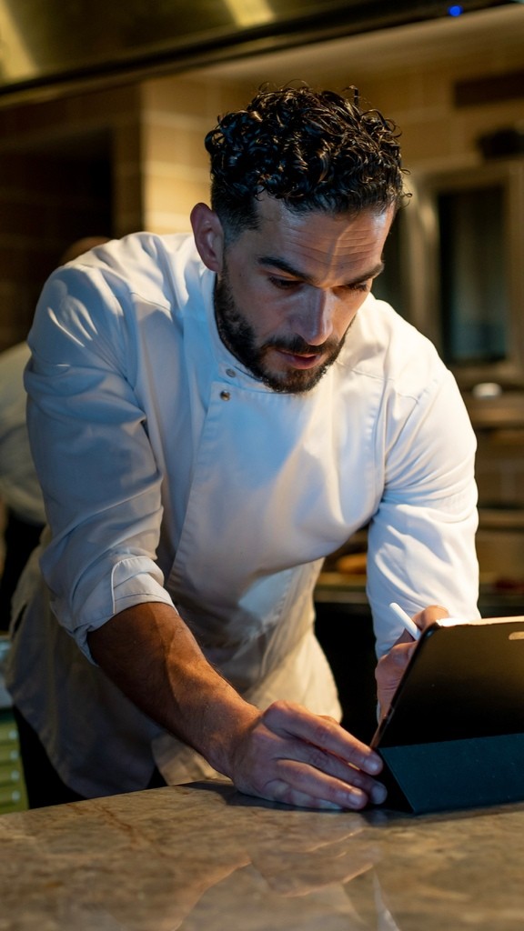 Jozef Youssef in a white chef’s jacket, concentrating on a tablet at a kitchen worktop