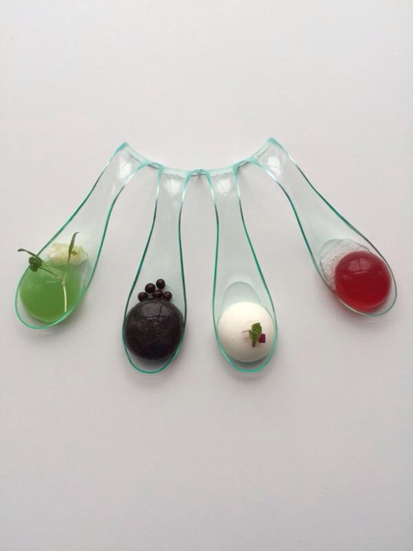 Four transparent tasting spoons with green, dark, white and red foods on a pale background
