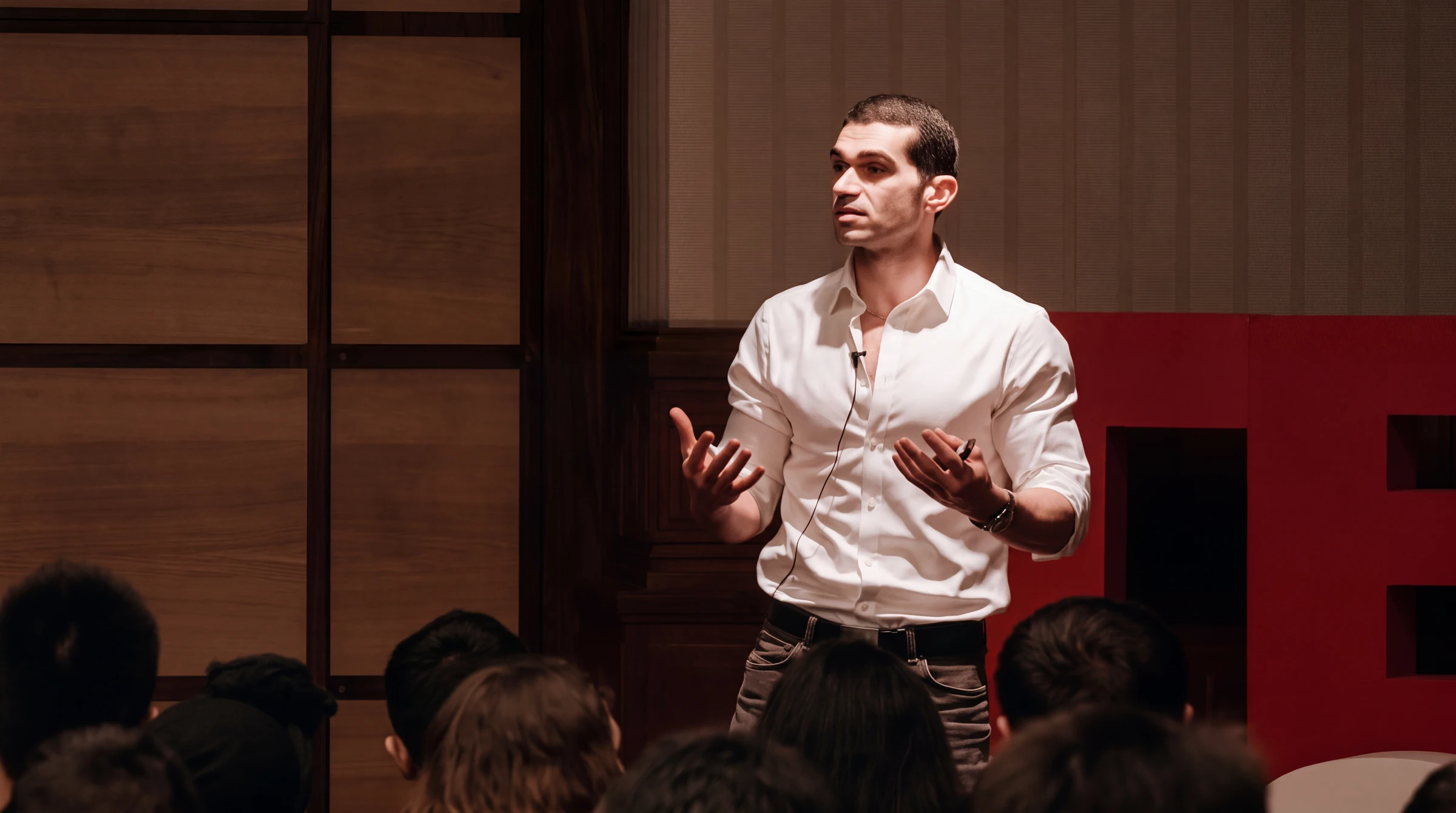 Jozef Youssef giving a TEDx talk in a white shirt, addressing an audience from a wood-panelled stage